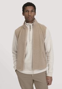 Beige Fleece-Weste mit hohem Kragen und Frontreißverschluss, kombiniert mit einem hellgrauen Pullover. Verfügt über Reißverschlusstaschen und eine weiche Textur.