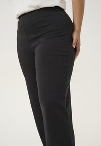 Pantalons noirs en matière lisse et extensible, présentant une taille haute et une coupe ajustée le long des jambes. Pas de motifs ni de quincaillerie visibles.