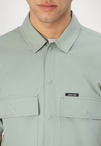 Light green button-up shirt with a collar, two chest pockets, and a black label on the left pocket. Made of textured fabric.