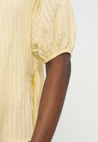 Χέρι ατόμου που φοράει υφασμάτινη ανοιχτοκίτρινη μπλούζα με φουσκωτά μανίκια κοντά με λάστιχο στις μανσέτες.