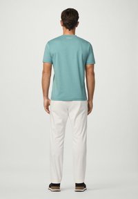 Muž stojící zády, oblečený v tyrkysovém tričku s krátkým rukávem, bílých kalhotách a černých teniskách s hnědými podrážkami na jednobarevném pozadí.