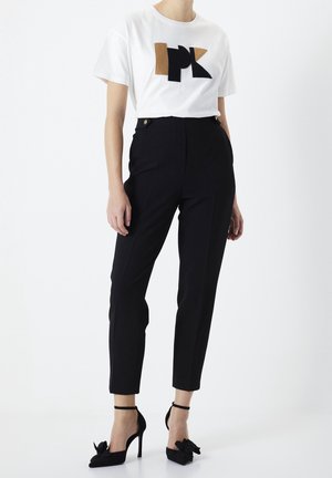 Femme portant un pantalon slim noir taille haute, un top blanc manches courtes avec un motif géométrique marron et noir, et des escarpins noirs avec brides à la cheville.