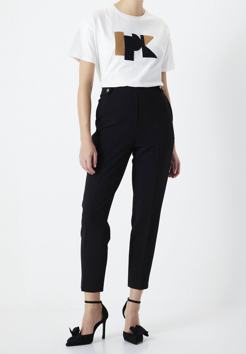Femme portant un pantalon slim noir taille haute, un top blanc manches courtes avec un motif géométrique marron et noir, et des escarpins noirs avec brides à la cheville.