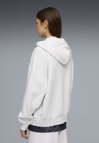 Γυναίκα με μακριά καστανά μαλλιά που φοράει άνετη, ανοιχτό γκρι φούτερ με κουκούλα και ταιριαστά παντελόνια φόρμας, στέκεται πλευρικά σε φόντο με γκρι διαβάθμιση.