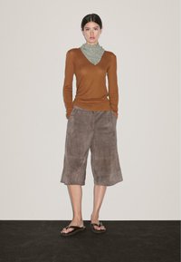 Woman standing against a plain wall wearing a brown long-sleeve top, gray scarf, loose gray knee-length shorts, and black flip-flops.