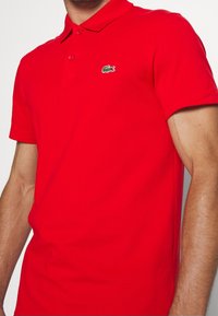 Červená polo košile s texturovanou tkaninou, krátkými rukávy a zapínáním na tři knoflíky. Na levé straně hrudi je zelené logo Lacoste.