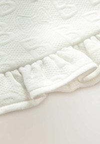Primer plano de una tela blanca texturizada con un borde rizado. Presenta un sutil patrón en relieve y una textura suave y lisa.