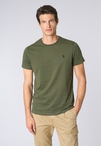 T-shirt di cotone verde oliva con collo rotondo, maniche corte e piccolo logo blu navy sul petto. Texture liscia. Abbinato a pantaloni corti khaki.