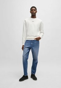 Valkoinen huppari, jossa on logo rinnassa, yhdistettynä sinisiin farkkuihin ja mustiin lenkkareihin. Slim fit -malleja, pitkät hihnat ja klassinen muotoilu.