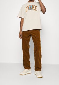 Béžové oversized tričko s grafikou "EVOKE", spárované s hnědými kordovými kalhotami a béžovými teniskami s texturovanou podrážkou.