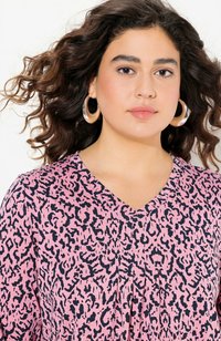 Roze top met een V-halsontwerp met een zwart luipaardpatroon. Model draagt grote hoopoorbellen. Zachte stof met een gestructureerde uitstraling.