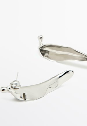 Pár lesklých náušnic z nepravidelného stříbrného kovu s puzetovými zapínáními na bílém pozadí.