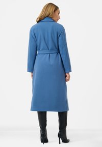 Cappotto lungo con cintura in tessuto blu testurizzato, con colletto a rever, caratterizzato da un design di media lunghezza e stivali neri al ginocchio.