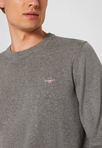 Man wearing a grey knit sweater with a small embroidered red and white shield logo on the left chest.