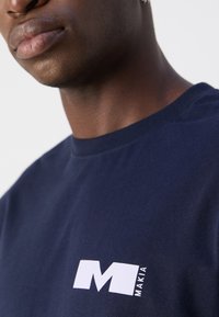 Námořnická modrá bavlněná tričko s bílým logem na levé hrudi, kulatým výstřihem a krátkými rukávy, s hladkým povrchem a ležérním designem.