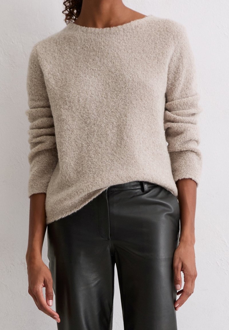 Pull beige texturé à manches longues associé à un pantalon en cuir noir, porté par une personne debout contre un mur clair uni.