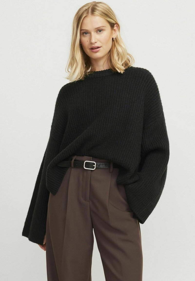 Un suéter negro de gran tamaño con textura acanalada que presenta mangas anchas y un cuello redondo, combinado con pantalones marrones de talle alto y un cinturón negro.
