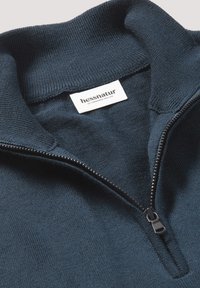Dunkelblauer Pullover mit hohem Kragen und halbem Reißverschluss. Weiche Stricktextur, versehen mit einem gewebten Etikett mit "hessnatur" und Gründungsdatum.