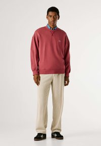 Sudadera color burdeos con puños acanalados y cuello redondo, combinada con pantalones acanalados beige. El conjunto incluye zapatillas negras informales con detalles en blanco.