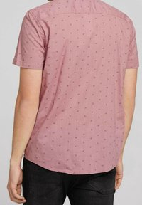 Camicia a maniche corte color rosa chiaro con motivi di aeroplani di carta blu scuro sparsi, realizzata in tessuto strutturato. Orlo arrotondato e dettaglio sulla spalla posteriore.