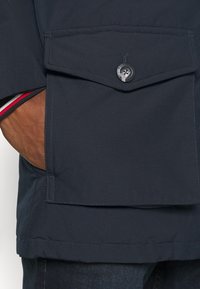 Tmavě modrá bunda s velkou klopovou kapsou, zaobleným designem, jemným prošíváním a zapínáním na knoflíky s logem.