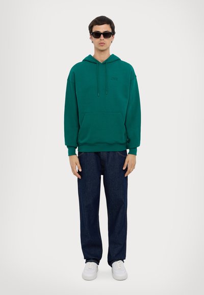 Teal green hoodie with a front pocket, drawstring hood, and logo detail. Paired with loose dark denim jeans and white sneakers.