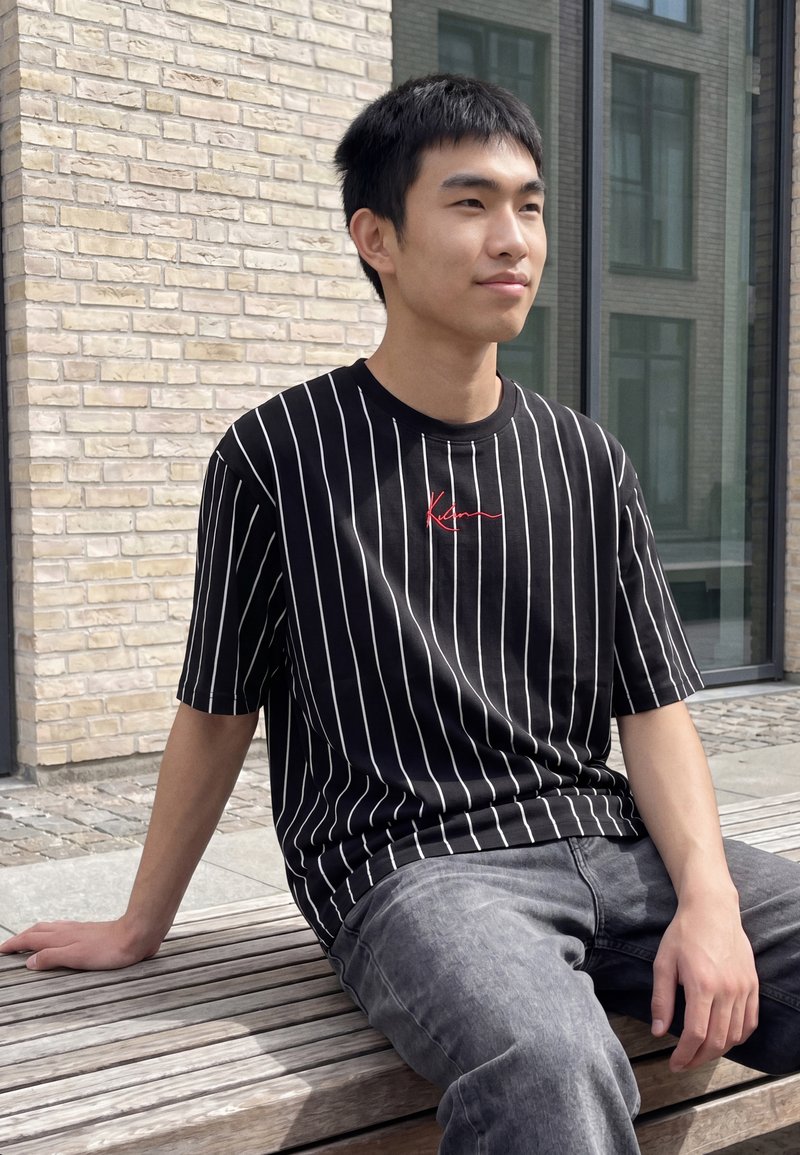 Joven con camiseta de rayas negras y blancas sentado en un banco de madera junto a una pared de ladrillos y una gran ventana.