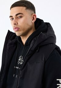 Sort puffer vest med hætte over en sort hoodie, der har et broderet logo og snoretræk detaljer. Glat stof og strømlinet design.