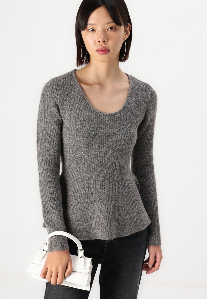 Maglione grigio a coste con scollatura a barchetta, maniche lunghe e orlo svasato, abbinato a una piccola borsa bianca.