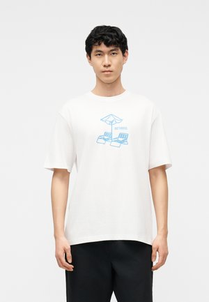 Nuori mies valkoisessa T-paidassa, jossa on sininen kuva kahdesta aurinkotuolista varjon alla ja sana "ELÄKELÄINEN".