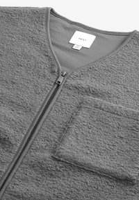 Veste zippée en polaire grise avec une surface texturée, col en V, et une poche poitrine. Elle possède une doublure lisse et une fermeture éclair argentée.