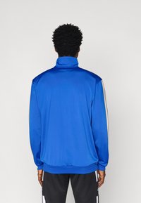 Μπλε σακάκι προπόνησης με ψηλό γιακά, λεία υφή, που διαθέτει κρεμ λωρίδες κατά μήκος των ώμων. Σχεδιασμένο για αθλητική χρήση, με μακριά μανίκια.