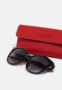 Black oversized sunglasses with gradient lenses beside a red leather case embossed with "GUESS." Case features a fold-over design.