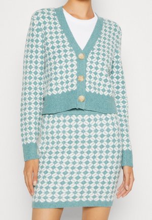 Femme portant un cardigan en maille assorti turquoise et blanc avec trois boutons et une jupe ajustée.