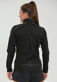 Zwarte sportjacket met hoge kraag, lange mouwen, mesh zijpanelen en reflecterende accenten. Voorzien van een grafiek van een hartslaglijn op de achterkant.