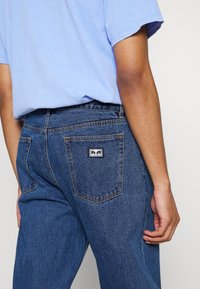 Dark blue denim jeans feature a high waist, straight leg design, visible stitching, and a patch logo on the back pocket.