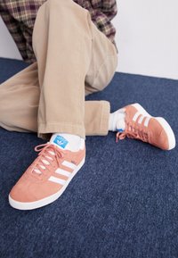 Sneakers in suede rosa con strisce e lacci bianchi, caratterizzati da un'etichetta blu, indossati con pantaloni beige su una superficie di tappeto blu.