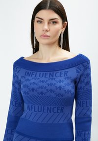 Top blu, a coste, con scollatura off-shoulder. Presenta un motivo testuale in rilievo e texture a contrasto. Orecchini luccicanti completano l'outfit.