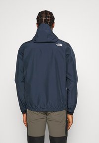 Giacca impermeabile blu con cappuccio, orlo elastico e polsini. Presenta un logo bianco sulla parte superiore della schiena. Tessuto liscio con finitura opaca.