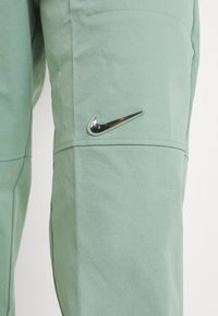 Ljusgröna byxor med en glänsande svart Nike swoosh-logotyp på låret mot en vit bakgrund.