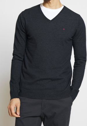 Mann trägt einen dunkelgrauen V-Ausschnitt-Pullover über einem weißen T-Shirt und dunkle Hose, steht mit einer Hand in der Tasche vor einem einfarbigen Hintergrund.
