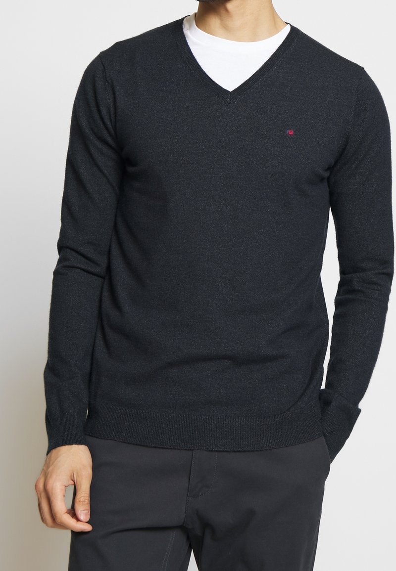 Homme portant un pull col en V gris foncé par-dessus un t-shirt blanc et un pantalon foncé, debout avec une main dans la poche sur un fond uni.