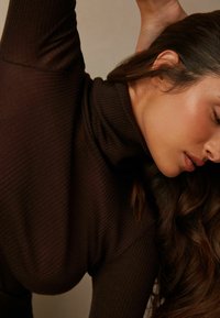 Hnědý žebrovaný rolák s těsným střihem, vyrobený z texturované látky, s vysokým límcem, částečně zakrytý dlouhými vlasy.