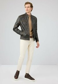 Bruine leren jas met een staande kraag, ritszakken, gecombineerd met een lichtbeige polo, crèmekleurige broek en donkerbruine sneakers.