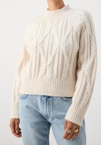 Pull crème en maille torsadée avec poignets et ourlet côtelés, porté avec un jean taille haute bleu clair, mains détendues le long du corps avec des bagues visibles.