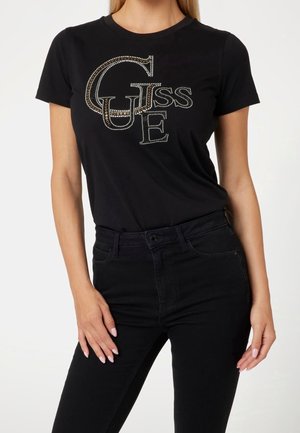 Kvinde iført en sort T-shirt med korte ærmer med dekorativt rhinestone "GUESS" logo og højtaljede sorte bukser, poserer foran en ensfarvet baggrund.