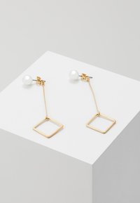 Goldene Tropfenohrringe mit einer weißen Perle als Stecker und einem geometrischen quadratischen Anhänger, der an einer feinen Kette hängt.