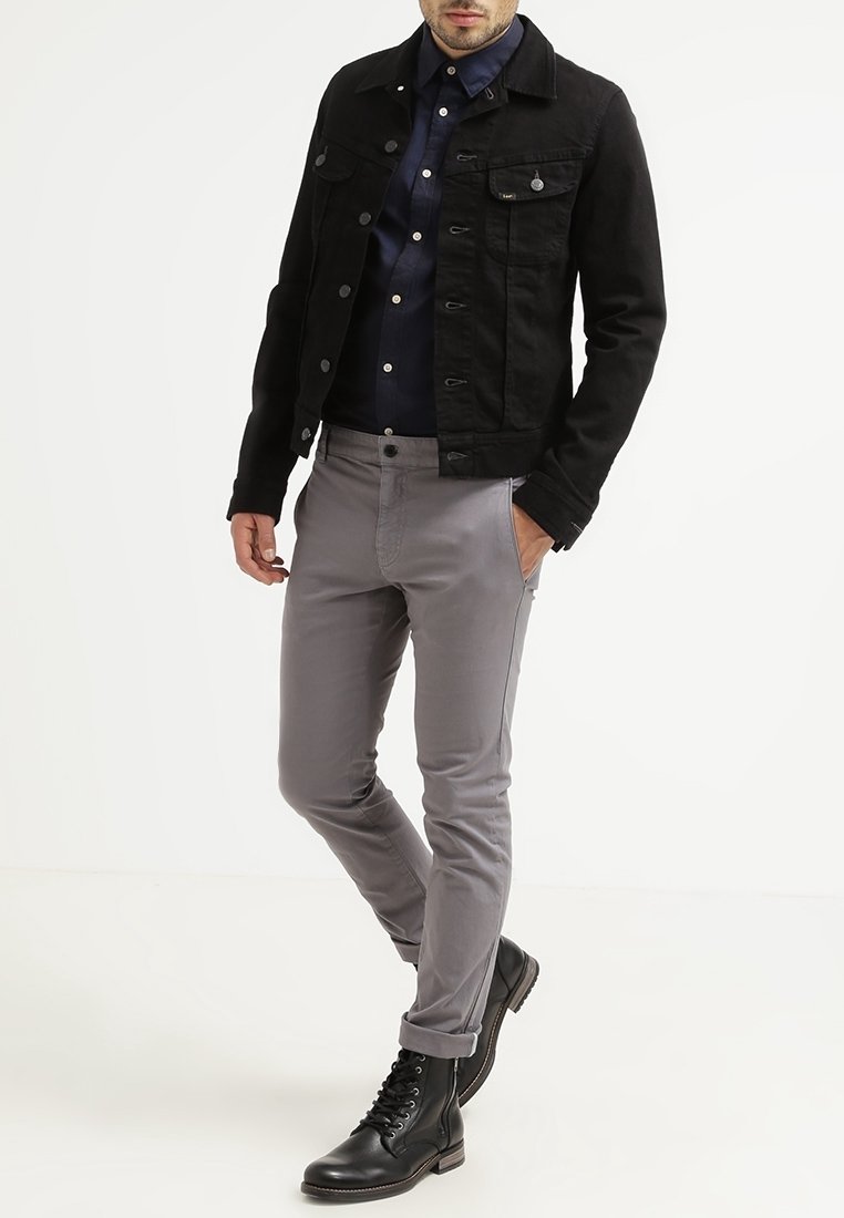 Homme portant une veste en jean noire, une chemise bleu marine, un pantalon slim gris et des bottes noires à lacets, une main dans la poche.
