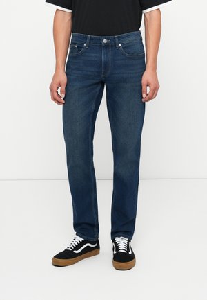 Dark blue denim jeans with a straight leg fit, featuring five pockets, a metal button fastening, and stitching details. Worn with black sneakers.