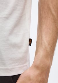 Κοντινό πλάνο στο τελείωμα ενός ανοιχτόχρωμου πουκαμίσου με μια μικρή μαύρη υφασμάτινη ετικέτα με την επιγραφή «BOSS» πορτοκαλί σε πλευρά του βραχίονα ενός ατόμου.